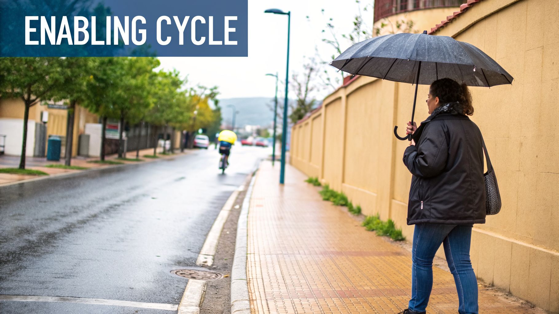 A person with an umbrella walks on a wet sidewalk next to a wall, with a cyclist in the distance.