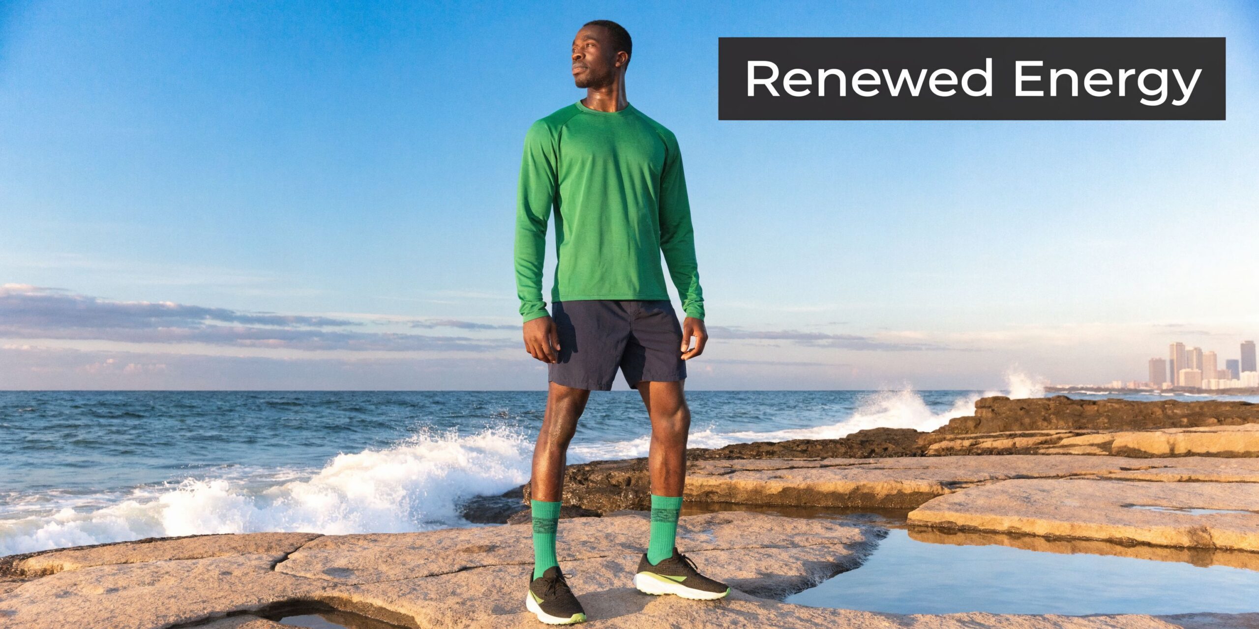 A man wearing a green long-sleeved shirt and shorts standing on a rocky beach by the ocean.