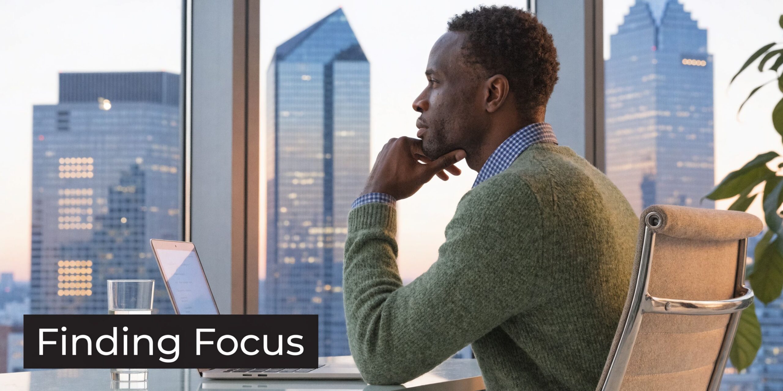 A professional man sitting at a desk by a window looking out at city skyscrapers while contemplating.