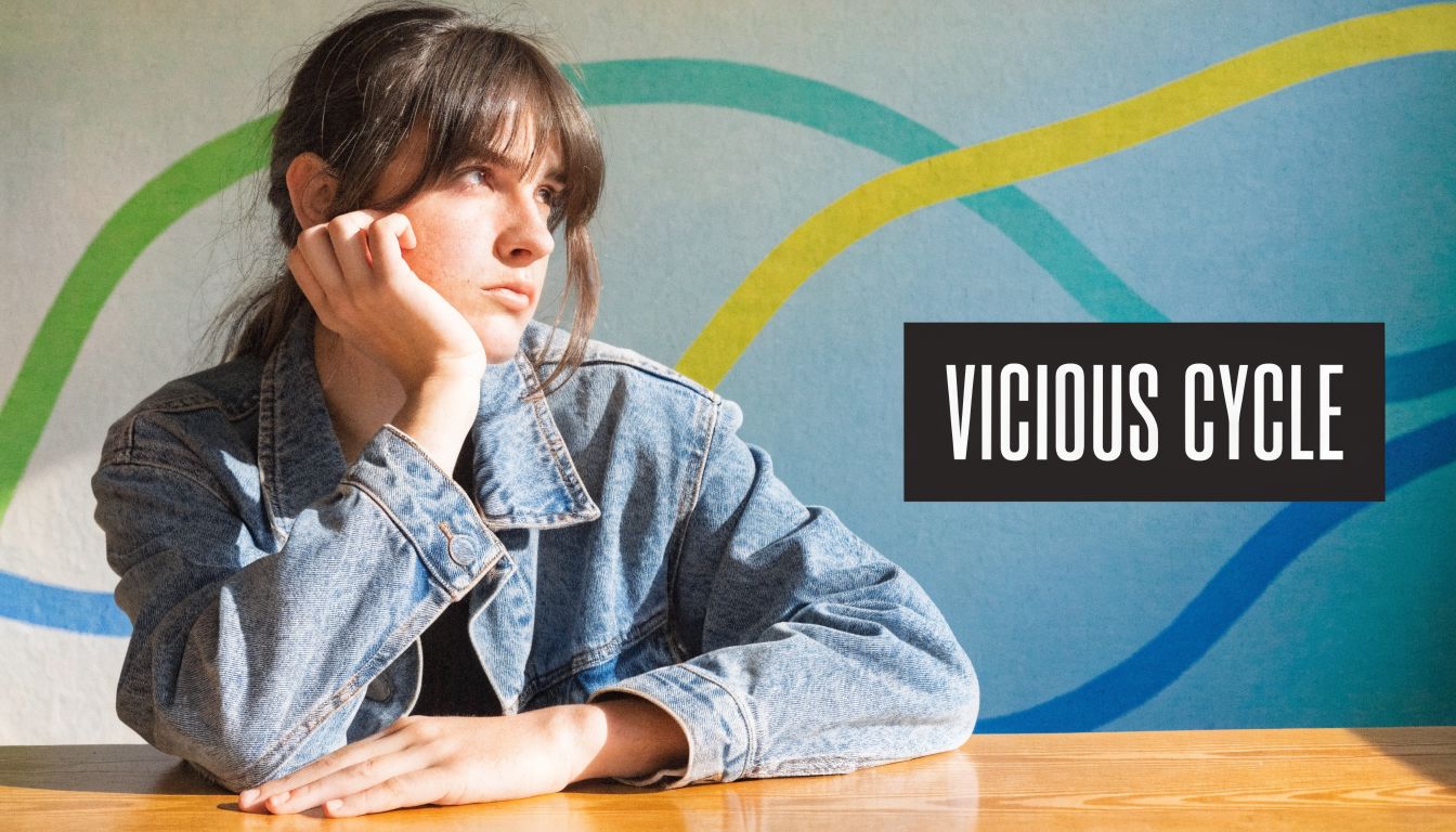 A pensive young woman in a denim jacket rests her head on her hand against a mural.